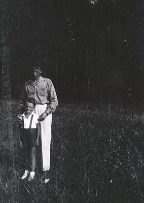 A mid-20th century black-and-white portrait of an adult holding a young child outdoors in overgrown grass. The adult wears a ...