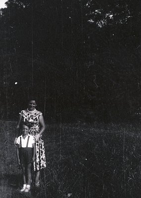 Black-and-white portrait of a woman and child in a grassy outdoor setting, likely 1950s–1960s. Woman wears a leopard-print dr...