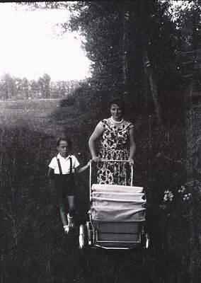 Black-and-white photo of a woman pushing a child in a wheeled cart along a rural path, likely mid-20th century. Floral dress ...