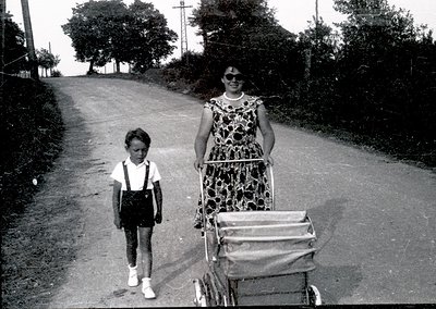 Black-and-white street scene featuring a woman in a floral dress and sunglasses pushing a vintage pram, accompanied by a youn...