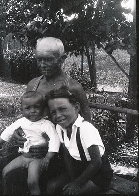 A mid-20th-century black-and-white portrait of a generational family in an outdoor setting. An elderly man in a striped shirt...