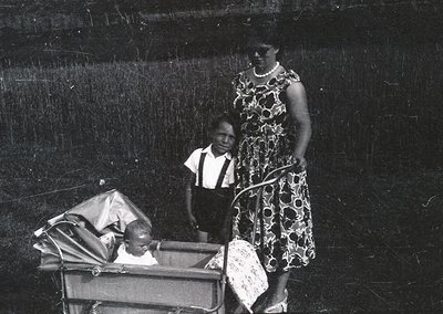 Vintage black-and-white photo of a woman in a floral dress and suspenders, pushing a baby carriage with a child seated inside...