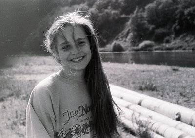 Black-and-white candid of a smiling child outdoors, wet hair framing face with sunlight rays creating lens flare. "Jay Na" gr...