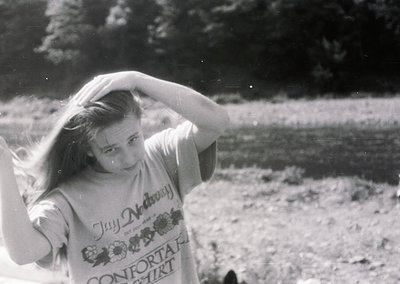 Vintage black-and-white photo of a young woman in a "Jay McDermott Comfort" t-shirt, outdoors near a body of water. Her hair ...