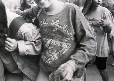 Black-and-white photo of three young women hugging outdoors, wearing 1970s-style sweatshirts with Sydney beach logos ("Coogee...