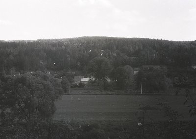 Rural landscape featuring dense forested hills in the background with scattered residential buildings and open fields below. ...