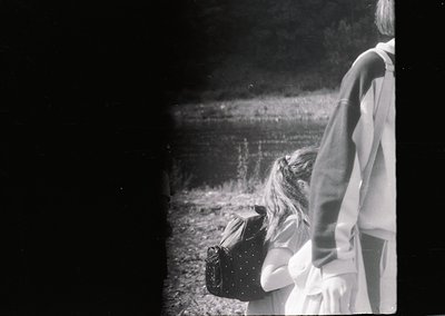 Vintage black-and-white photo of two children standing near a body of water, one holding a vintage suitcase. The scene sugges...
