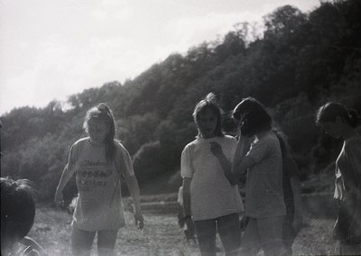 Vintage black-and-white photo of four young women in casual summer attire, likely 1970s, standing outdoors in a wooded area. ...