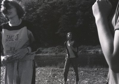 Black-and-white candid of three young people in outdoor setting, likely 1970s–1980s. Foreground: person in loose-fitting swea...