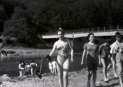 Vintage black-and-white photo of a group by a riverside in the 1960s–70s. Central figure wears a bikini with handwritten text...