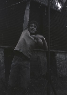 Vintage black-and-white photo of a young boy mid-jump, shirtless with rolled-up pants, against a dark, textured wall. Motion ...