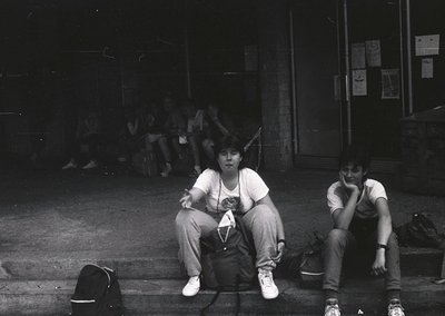 Black-and-white candid shot of two individuals seated on concrete steps in an urban setting, likely 1970s–1980s. The person o...