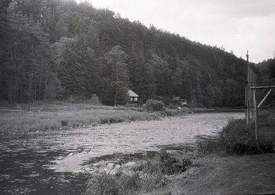 Black-and-white rural scene featuring a shallow river bordered by rocks and reeds, flanked by dense forest. Two modest wooden...