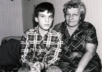 Black-and-white portrait of a young boy and elderly woman in matching plaid shirts, seated indoors. The woman’s floral-patter...
