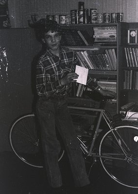 Black-and-white snapshot of a young person (likely 1970s) holding a folded paper in front of a bookshelf filled with canned g...