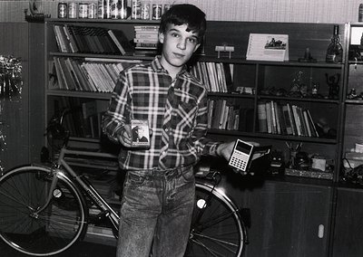 Mid-20th century boy poses indoors beside a vintage bicycle and early calculator. Bookshelves filled with hardcover volumes a...