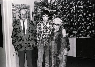 Intergenerational portrait featuring a man in a patterned blazer and glasses, a young boy in a plaid shirt, and an elderly wo...