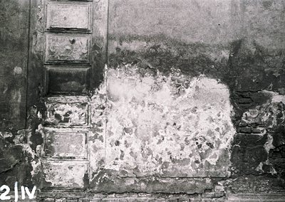 Black-and-white close-up of a damaged plaster wall with exposed brickwork and crumbling debris, likely from a bomb or artille...