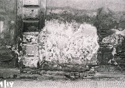 War-damaged brick facade with exposed plaster and mortar, likely post-WWII. Cobblestone street and partial stone archway visi...