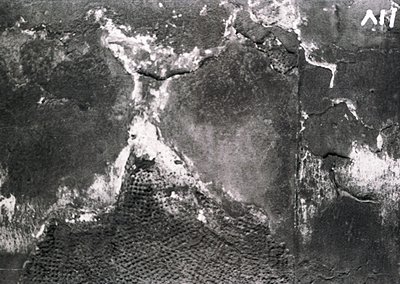 Aerial black-and-white photo of a coastal landscape showing eroded cliffs and a narrow beach. Distinctive rock formations and...