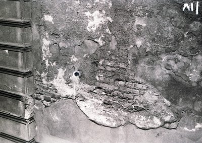 Close-up of severely damaged plaster and exposed brickwork, likely from a bomb or artillery strike. The jagged, uneven surfac...