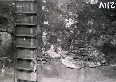 Black-and-white close-up of weathered concrete wall showing severe structural damage—exposed rebar and cracked plaster. Likel...