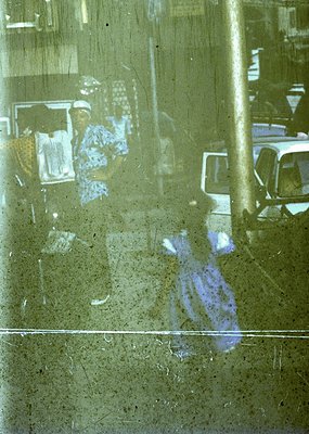 Reflection of a 1970s-era bus station interior: blurred passengers seated on patterned upholstery, a white van parked outside...