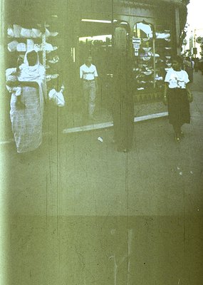 Reflection of 1970s urban street scene in wet pavement, showing blurred figures in casual attire (white shirts, dark pants) a...