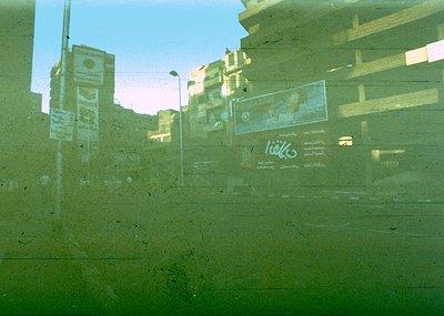 Reflection of urban street scene through a fogged or scratched window. Visible: faded billboards (Pepsi, Arabic script), mult...