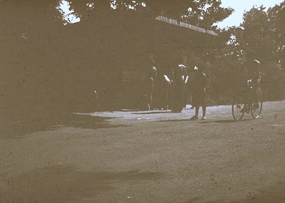 Vintage sepia-toned street scene showing three figures in early 20th-century attire—long coats, hats, and bicycles—walking ne...
