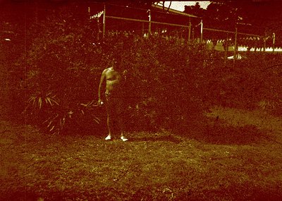 Vintage sepia-toned photo of a person mid-jump in a grassy area, surrounded by dense foliage. The individual wears a sleevele...