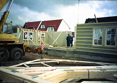 Mid-construction residential site featuring skeletal timber-frame homes ( ). Two workers in workwear inspect framing, with a ...