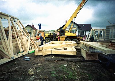 Construction site featuring a yellow ADHER crane lifting prefabricated wooden beams. Workers in blue uniforms oversee the ass...