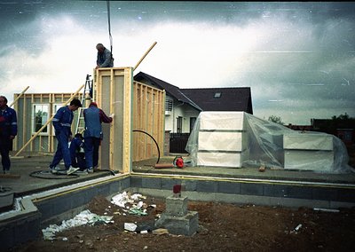 Construction crew assembling wooden frame on modern residential site, 1980s-1990s. Workers in blue overalls secure beams; par...