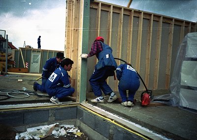 Three construction workers in matching blue uniforms with numbered tags assemble wooden framing on a rooftop. Snow and debris...