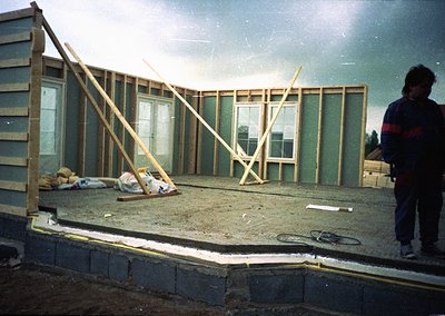Mid-stage construction of a single-story home, featuring green insulated panels and wooden framing. A worker in dark pants an...