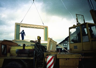 Construction worker hoists large wooden window frame via crane at industrial site, likely mid-20th century. Overcast skies an...