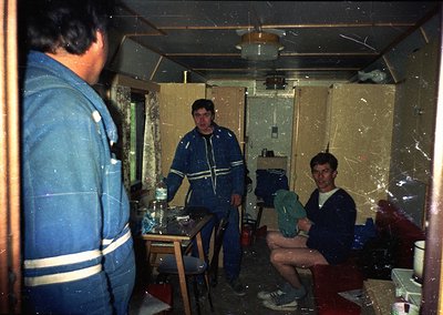 Indoor scene featuring three men in a utilitarian, dimly lit space resembling a ship’s or industrial cabin. The man on the le...