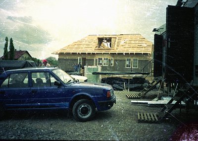 Vintage blue sedan parked beside a partially demolished single-story house under exposed roof framing. Construction debris an...
