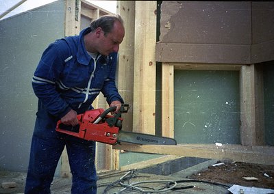 Man in 1970s-era workwear uses Husqvarna chainsaw on unfinished wooden frame, likely residential construction. Note vintage s...