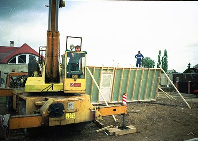 Construction site featuring a yellow hydraulic lift truck (model ) with a worker elevated in the bucket. Another worker stand...