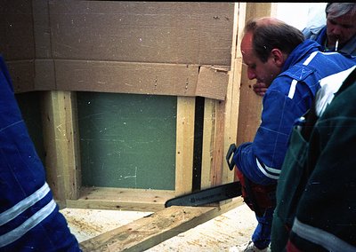 Construction workers in blue uniforms with reflective stripes examine a framed window opening in a partially built wooden str...