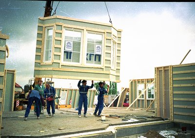 Four construction workers in matching overalls guide a prefab two-story unit via pulley system at a mid-century site. Light b...