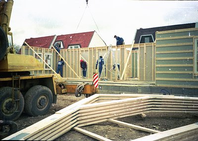 Construction crew erects prefab wooden panels using a crane on a residential site, mid-20th century. Uniforms suggest industr...