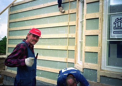 Two construction workers install prefab wall panels, 1970s-80s industrial housing era. Worker in red cap wields a tool, while...