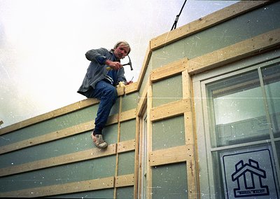 Mid-20th century construction worker securing plywood sheathing on a building frame. Overalls, tool belt, and safety-consciou...