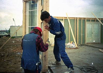 Two construction workers assemble prefab panels in a temporary site, likely 1980s–1990s Eastern Europe. Worker in red cap hol...