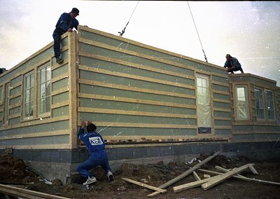 Pre-fabricated modular building construction in progress, featuring light green siding and white-framed windows. Workers in b...