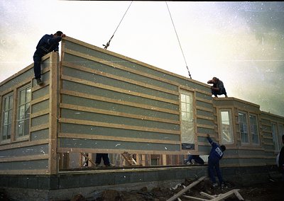 Mid-20th century construction crew hoists prefab wall panels using a crane. Workers in dark uniforms secure panels to a modul...