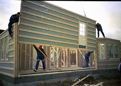 Mid-20th century construction crew framing a two-story wooden house, likely . Workers in blue coveralls and red caps secure h...